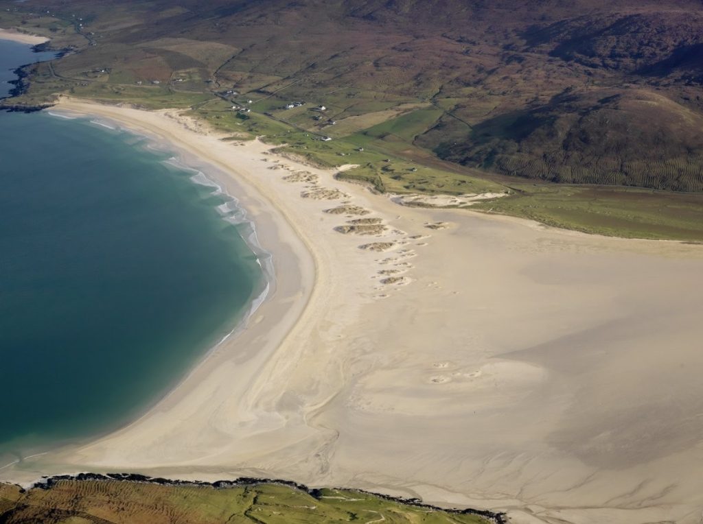 Obliques aerial photograph of a long white sand beach next to a green, grassy landscape. 