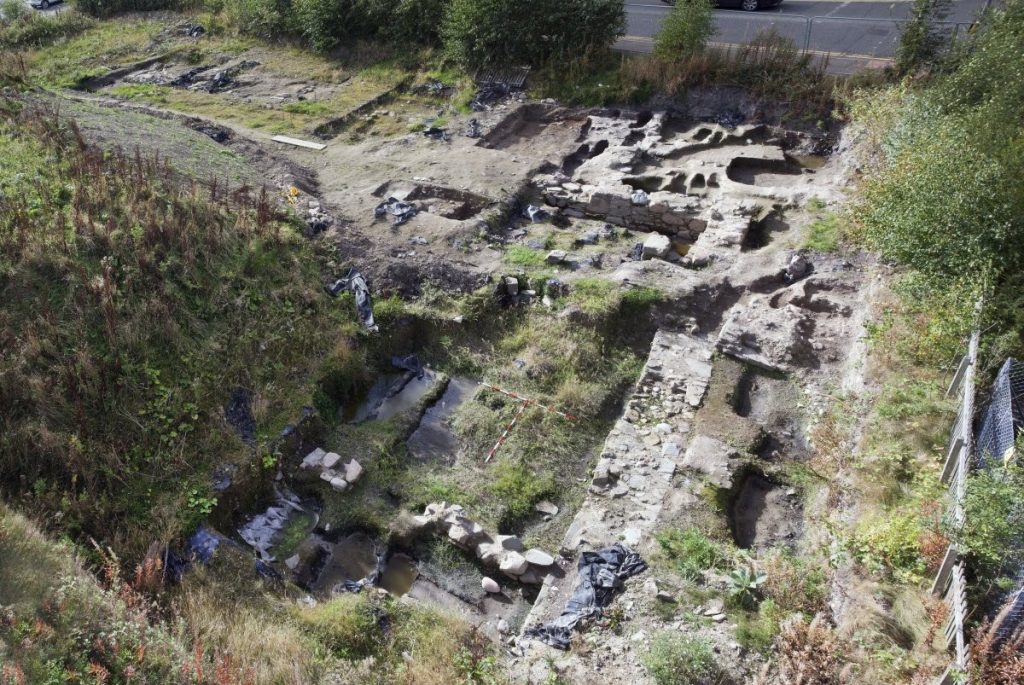 Oblique aerial view of a rectangular stone structure in ruin, surrounded by grass and overgrown weeds.