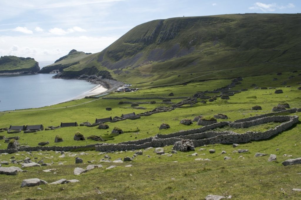 Landscape view over an abandoned village of houses and other stone buildings set in a green valley next to a large green hill. A body of water can be seen in the top left corner. 