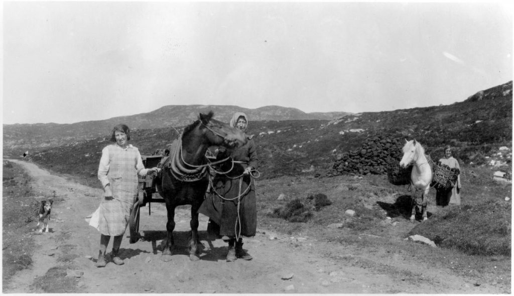 Black and white photgraph showing three women in 1930s dress. Two of them are standing on either side of a horse who is tied to a cart, and the other woman is standing next to a horse with two larbe baskets tied to its back. A dog stand nearby the women and horses. 