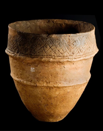 Photograph of a yellow-brown oval shaped urn with carved lines around the rim and a rounded base against a black background.
