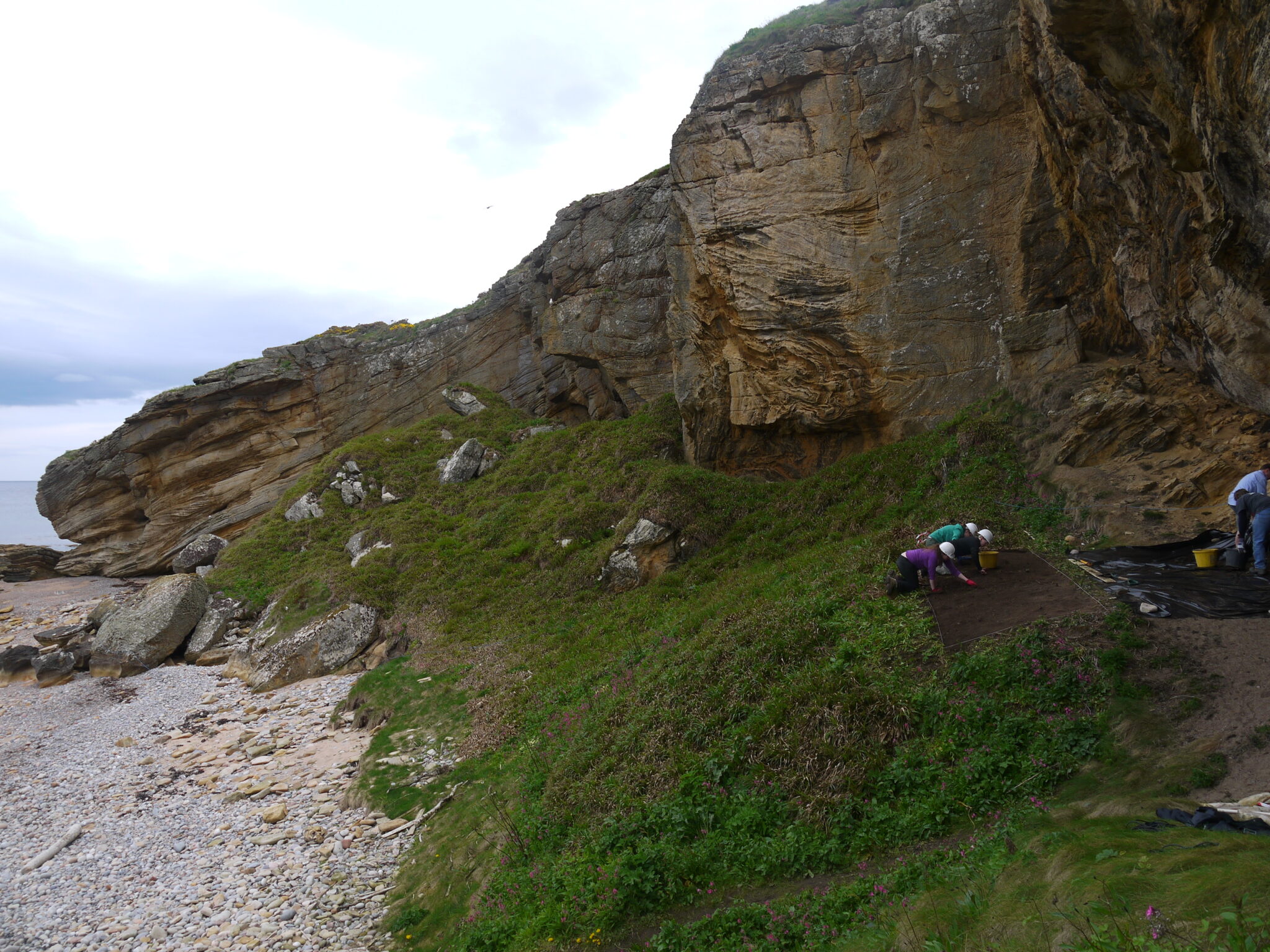 A wideshot of a cliffside on a grassy and stone covered beach. Three people can be seen next to the cliffside digging in a rectangular trench wearing white hardhats. 