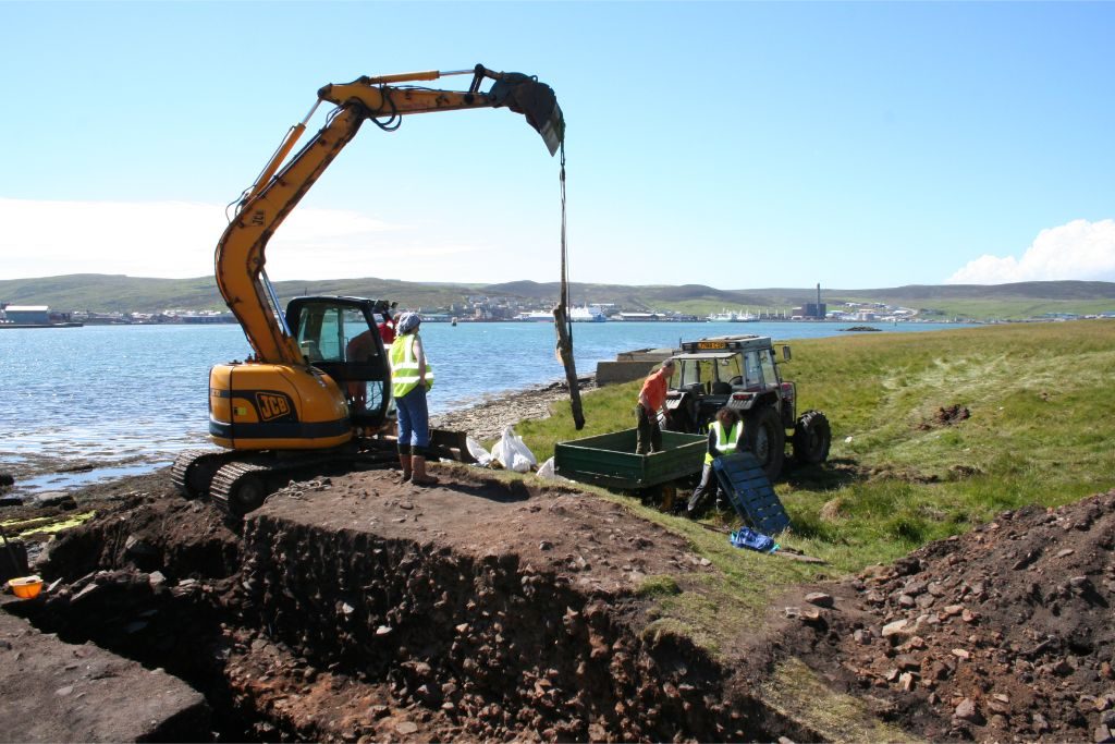 A large hydraulic machine with a lifting claw is placed next to an open excavation and in front of a large body of water. Four people are working nearby, lifting equipment or supervising the machinery. 