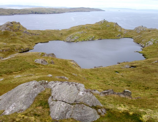 Oblique aerial view of a small lochan. An irregularly shaped body of water is surround by grass and heather, with a much larger body of water in the background. 
