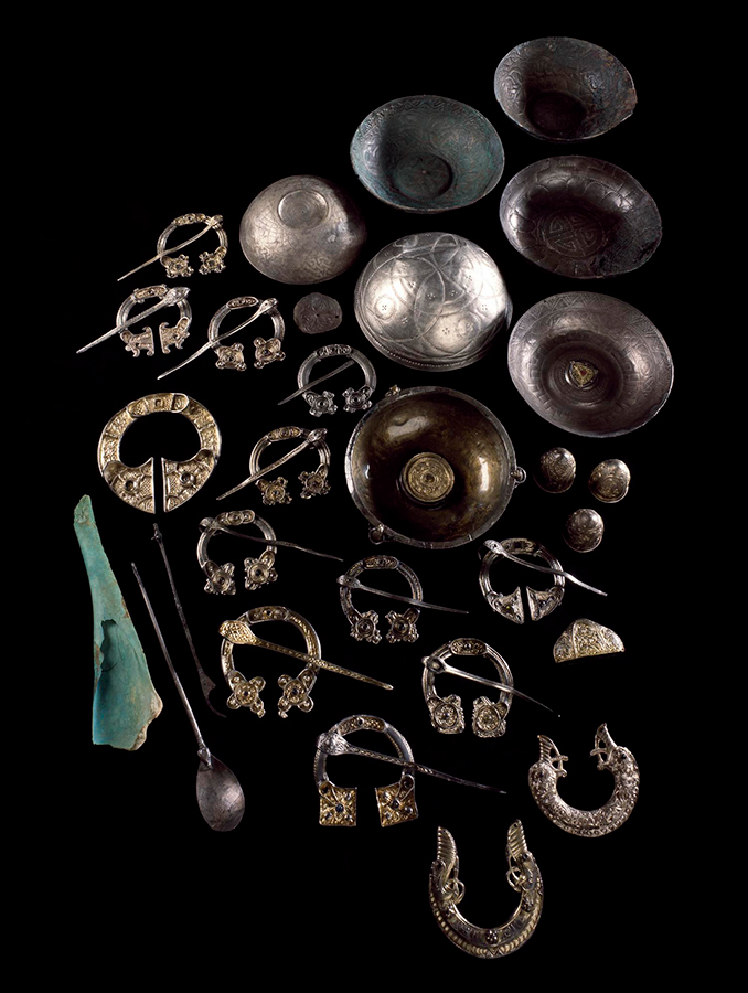 A collection of silver artefacts against a black background. The collection includes seven small bowls, fourteen semi-circular brooches and two spoons. 