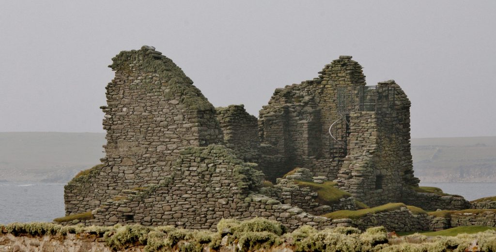 A ruined brown stone building with rectangular walls. A metal spiral staircase can be seen to the right side of the building, with a metal balcony at the top. The sea can be seen in the background. 