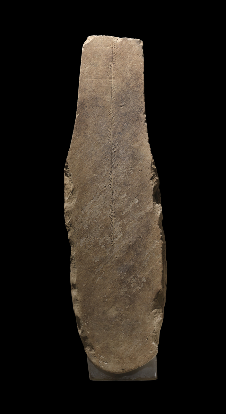 A long upright, sandstone slab against a black background. The stone has a clear strip of straight line carvings down the centre which resemble hashmarks. A faint cross can be seen in the top left corner. 