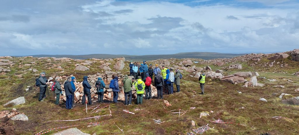 A group of 23 people, some wearing high vis vests, gather around a ruined stone structure in a grass and rock landscape. It is a cloudy day and most of the people have their hoods up. 