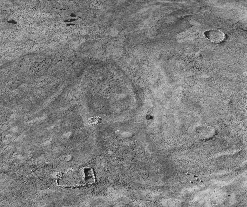 Black and white aerial view of a flat landscape with the remains of clear stone structures in various shapes spread across the image. Some are rectangular and others are round or oval. 