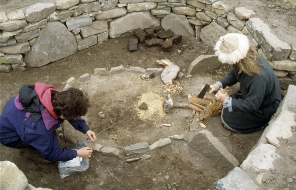 Two people are kneeling inside a stone structure with a stone lied hearth in the centre. One person is holding a piece of wood and an axe, and the other is unraveling wire. 
