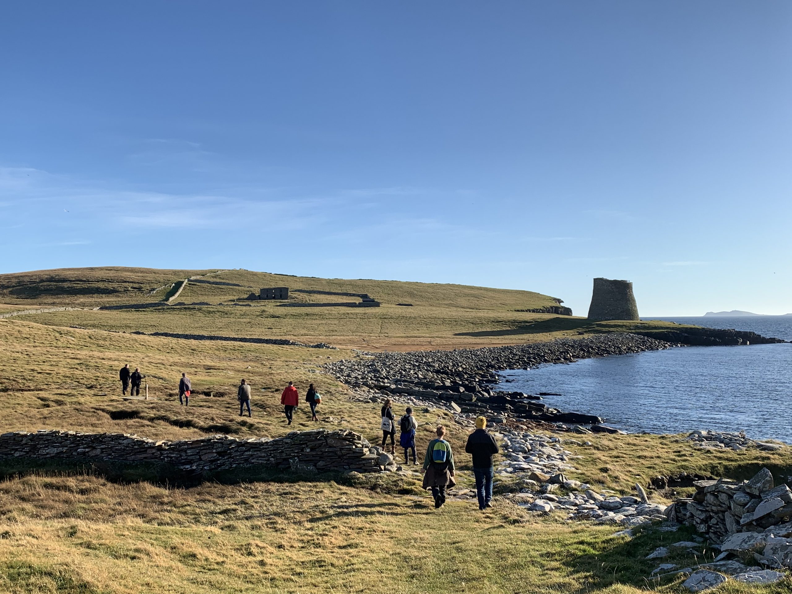 A group of eleven people wearing waterproof jackets walk along a coastline on grass towards a stone tower building. The Sky is blue and the water to the right of the image is reflecting the blue light.