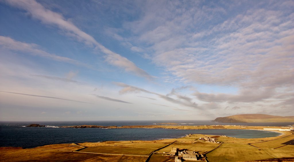 A wide angle view of a coastal landscape with grass-covered land and large inlets of blue water. The sky is blue and has strips of white and grey clouds. 