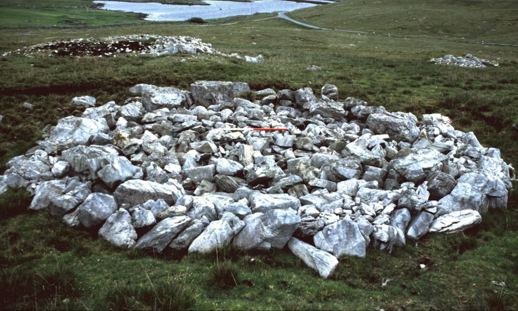 A large, flat, rounded pile of grey stones in a grassy hillside near a body of water. In the background, two more similar structure can be seen within the landscape. 