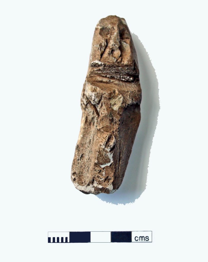 Photograph of a small, oblong, brown stone object with a face and arms carved into it using puncture marks and line carvings. The figure appears to be wearing a cloak.