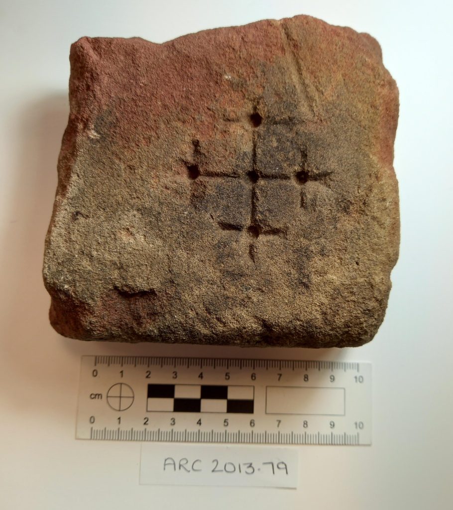 A square fragment of red-brown stone sits on a white background with a small ruler showing it is around 10cm wide. On the top is a cross made of deep punture holes and less prominent carved lines.