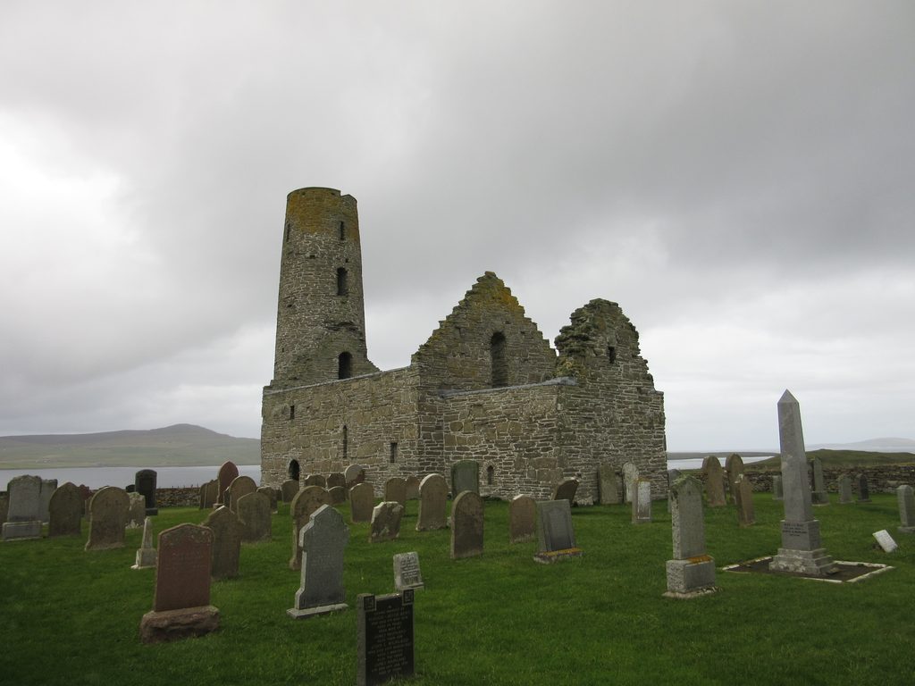 A ruined church building with a rounded tower on the left side, sits in a grassy graveyard with a body of water in the immediate background.