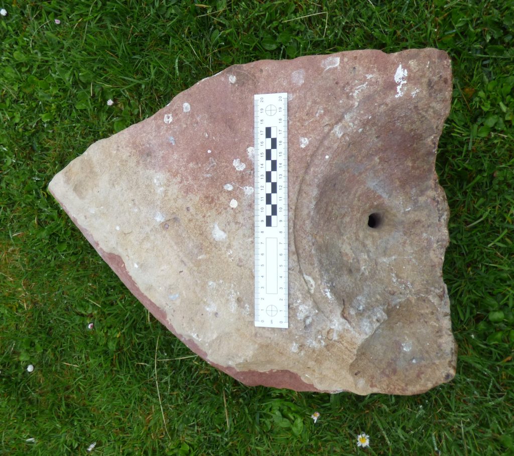A fragment of red stone in a pointed shape sits on grass with daisies growing. A ruler sits on top of the stone.