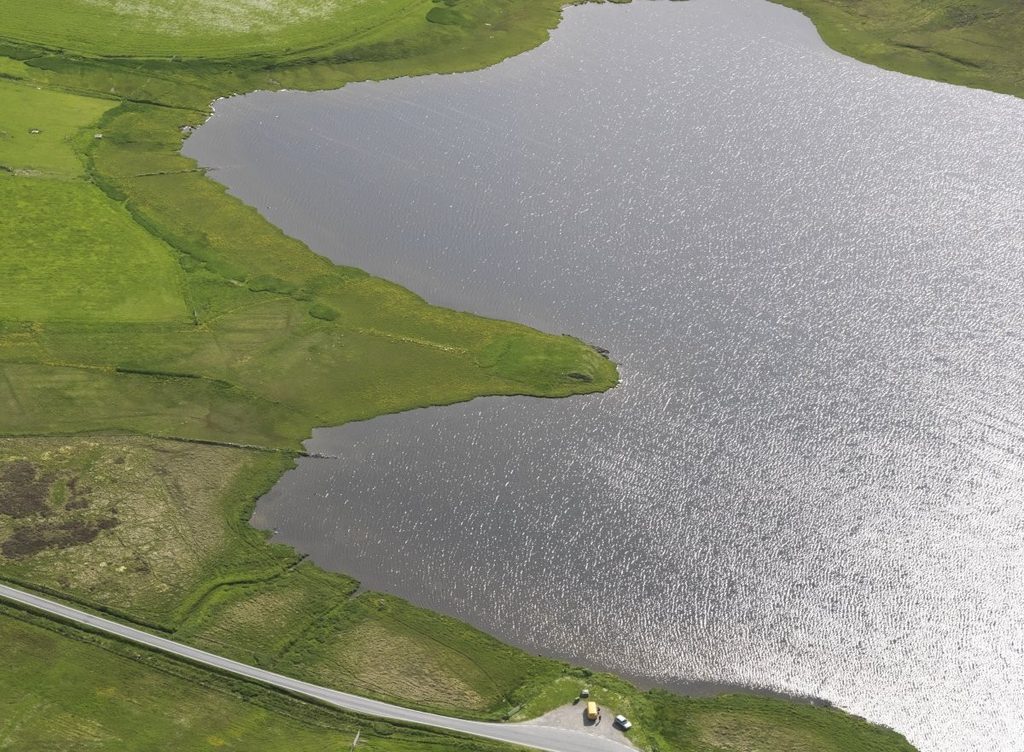 Aerial view of a grassy landscape with a large body of water to the right of the image. A long strip of land protrudes into the water, and the photo is focussed on this area.