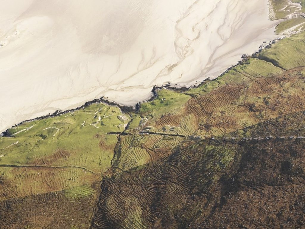 Aerial photograph of a green and brown landscape next to a white sandy beach. On the grass, clear lines are seen showing the remains of fields and their boudaries.