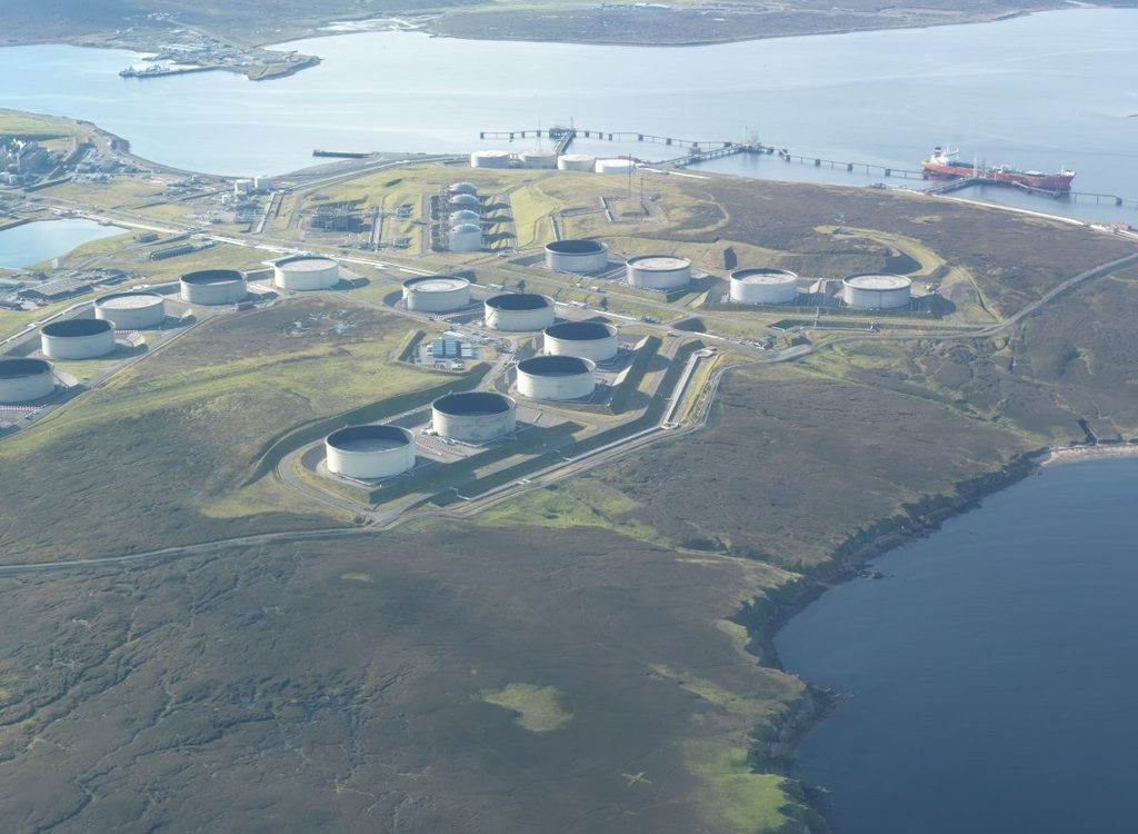 Oblique aerial view of fifteen cylindrical structures across a grassy, coastal landscape. The building are white and surrounded by concrete roads to connect them. In the background, a large bridge and sea vessel can be seen sitting next to the harbour. 