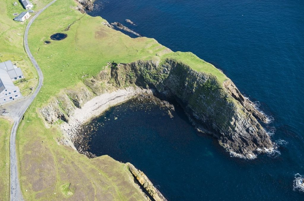 Aerial view of a cove surrounded by a grass covered cliffside. The cove has a sandy and stone covered beach at the bottom of the steep grassy cliff. The surrounding water is blue. 