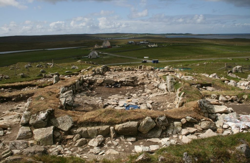 A rectangular stone sturcture with low drystone walls sits in a vast grassy landscape. The tops of the ruined walls are covered in turf and moss. 