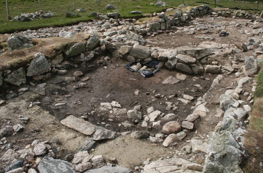 A large, rectangular structure under excavation. The building has square edges and long, drystone walls set into the ground. The building is surrounded by a grassy landscape. 