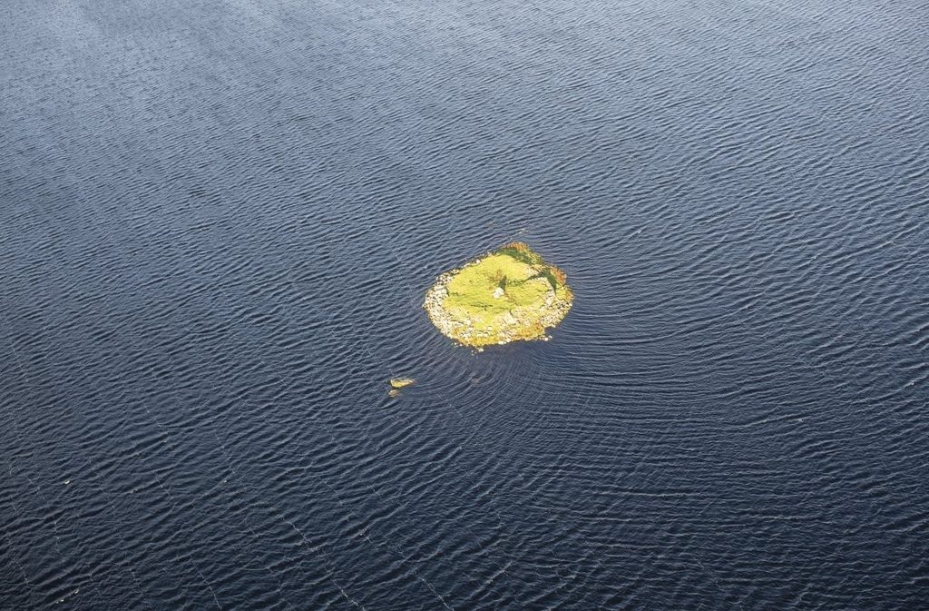 Oblique aerial photograph of a large body of water with a round island in the centre. The island is grass-covered, and a stone wall can be seen around the edge of the island. 