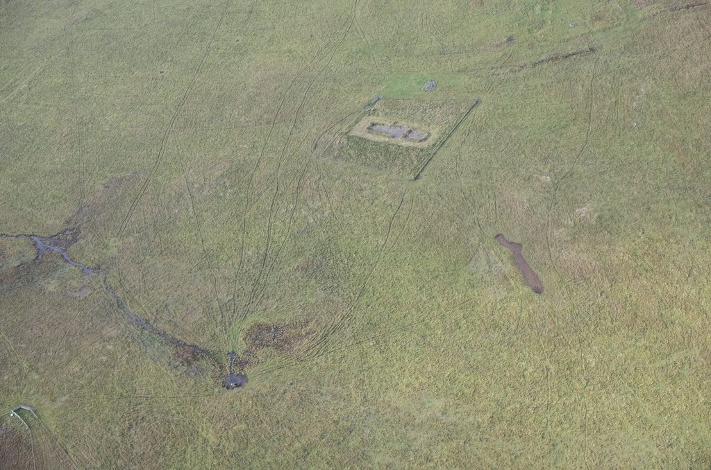 Oblique aerial view of a large grassy landscape showing archaeological features include a long, rectangular structur which is exposed and is grey against an otherwise green, grassy background. 