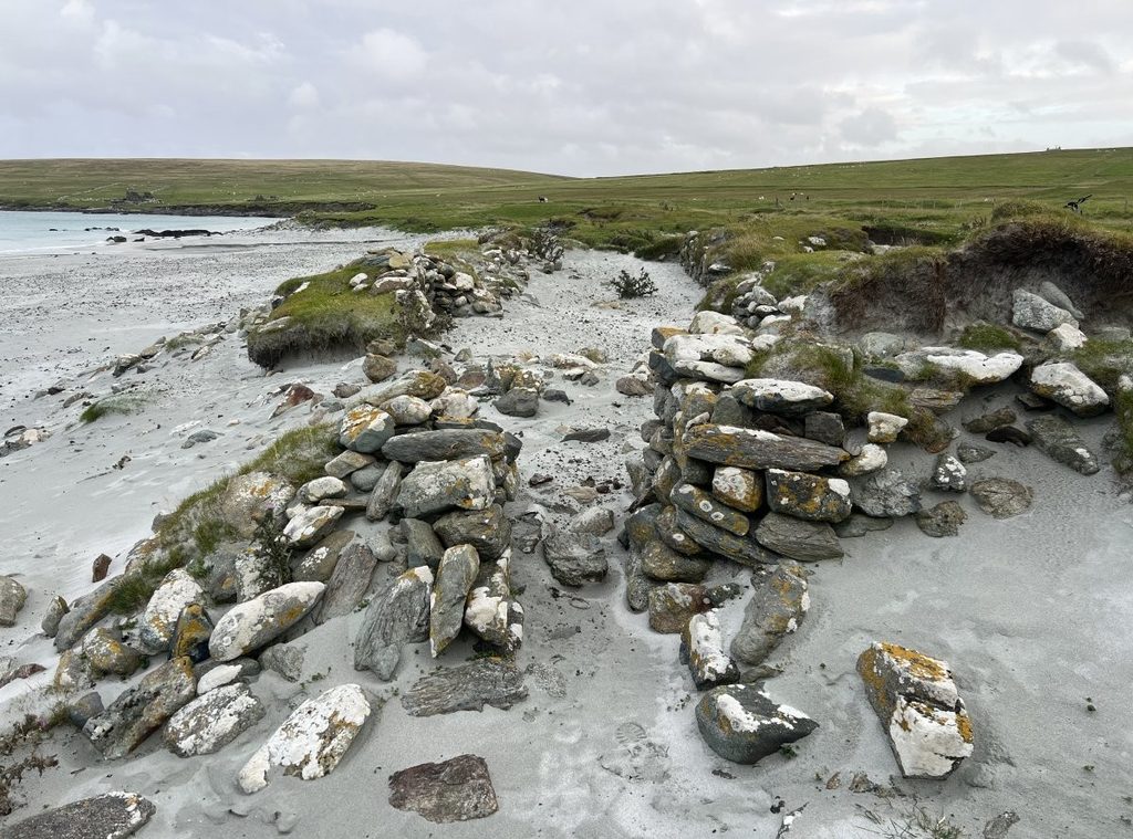 Photograph of a ruined stone longhouse on a sandy beach. The stones are placed in a long, rectangular shape, some of which are protruding from a grassy beach cliff. 