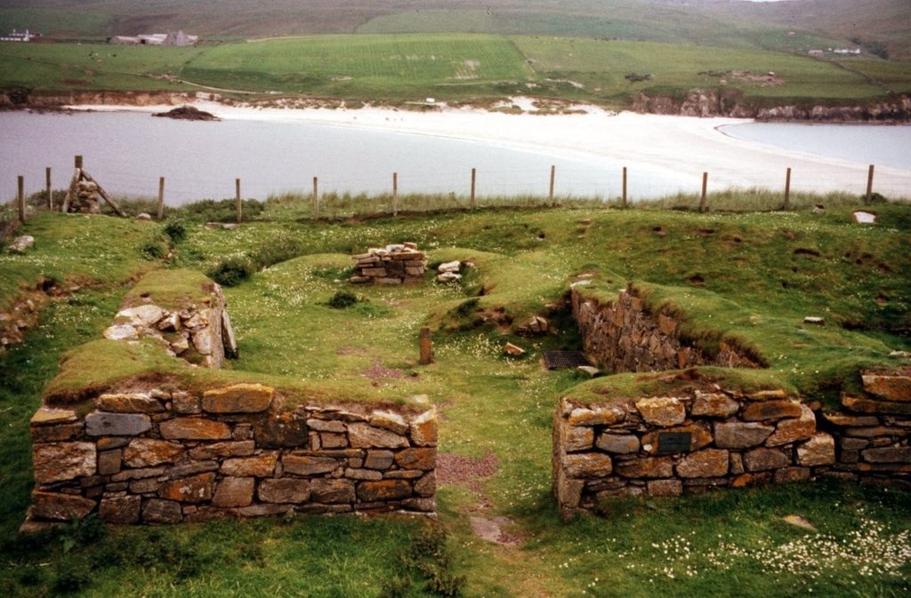 Rectangular stone structure with neat, drystone walls which are covered on the top with turf and moss. In the background, a white sand beach connects two areas of grassy land, with water on either side. 