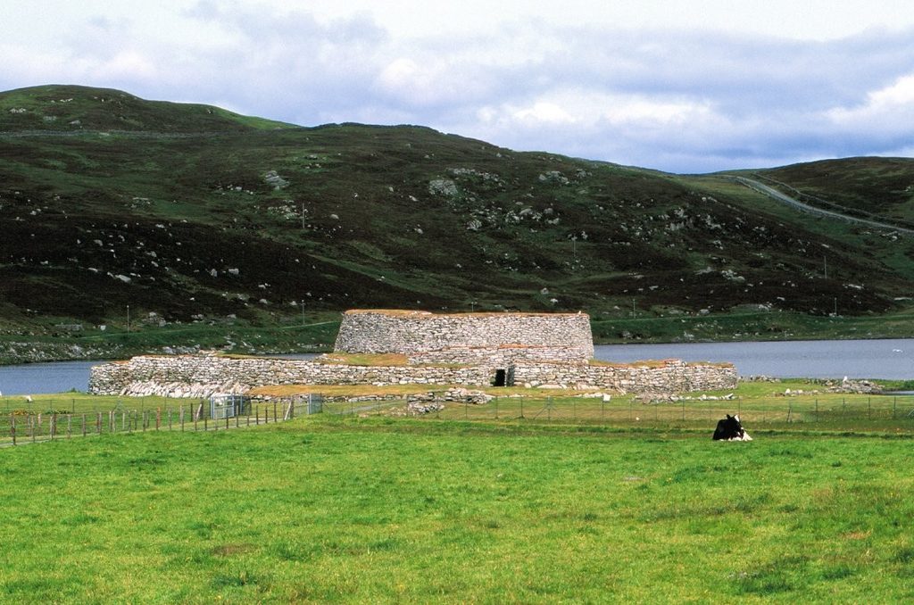 Image of a ruined stone tower, with a low drystone wall surrounding it. The settlement is on the edge of a grassy landscape, with water in the background of the photo. 