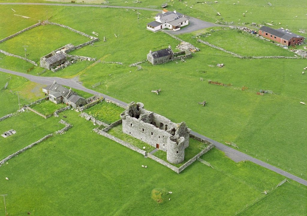 Oblique aerial photograph of a large grassy landscape, focussed on a grey stone ruin with no roof. A low rectangular wall in the same grey stone surrounds the castle-like structure. 