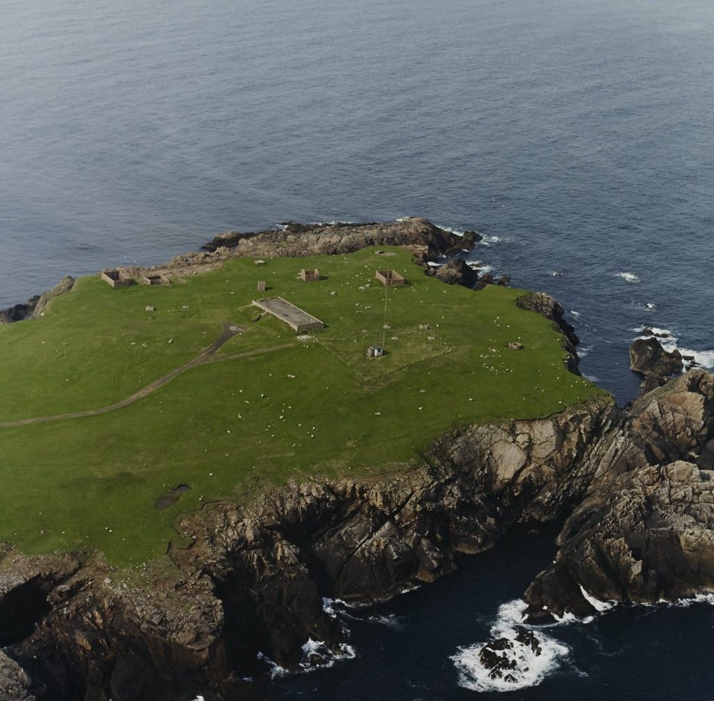 Aerial view of a small peninsular with steep, rocky cliffs and a flat, grass covered patch of land on top. On the grass, a rectangular building and tall metal rods can be seen from a distance. 