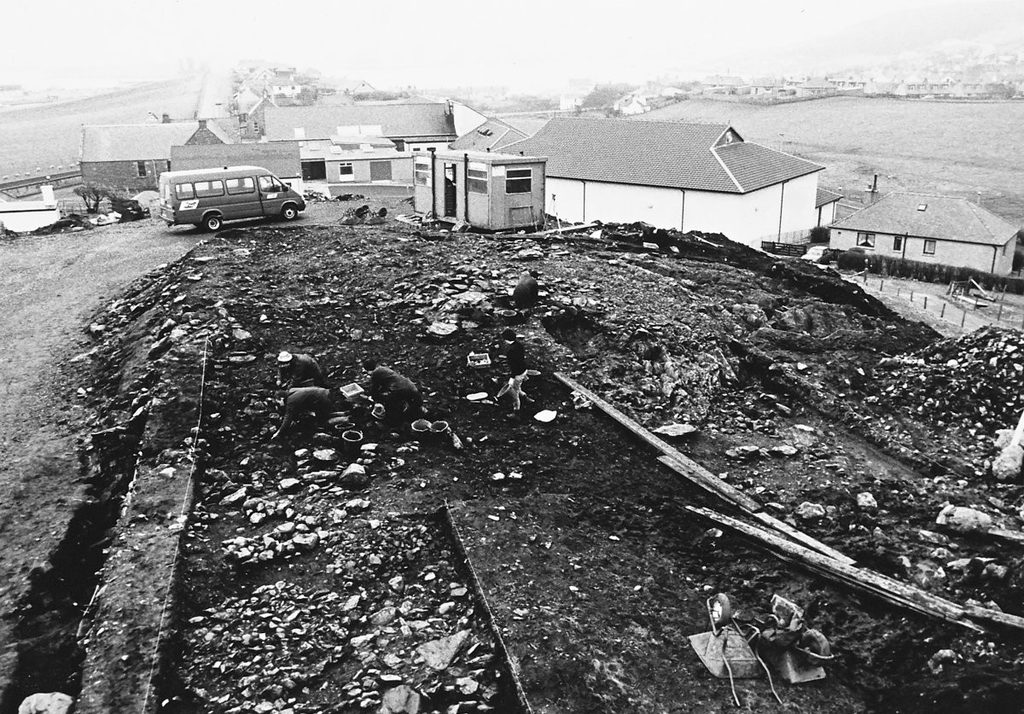 Black and white photograph of an open excavation, showing people crouching with trowels and buckets in a large, stony trench. A portable cabin and van can be seen in the background. 