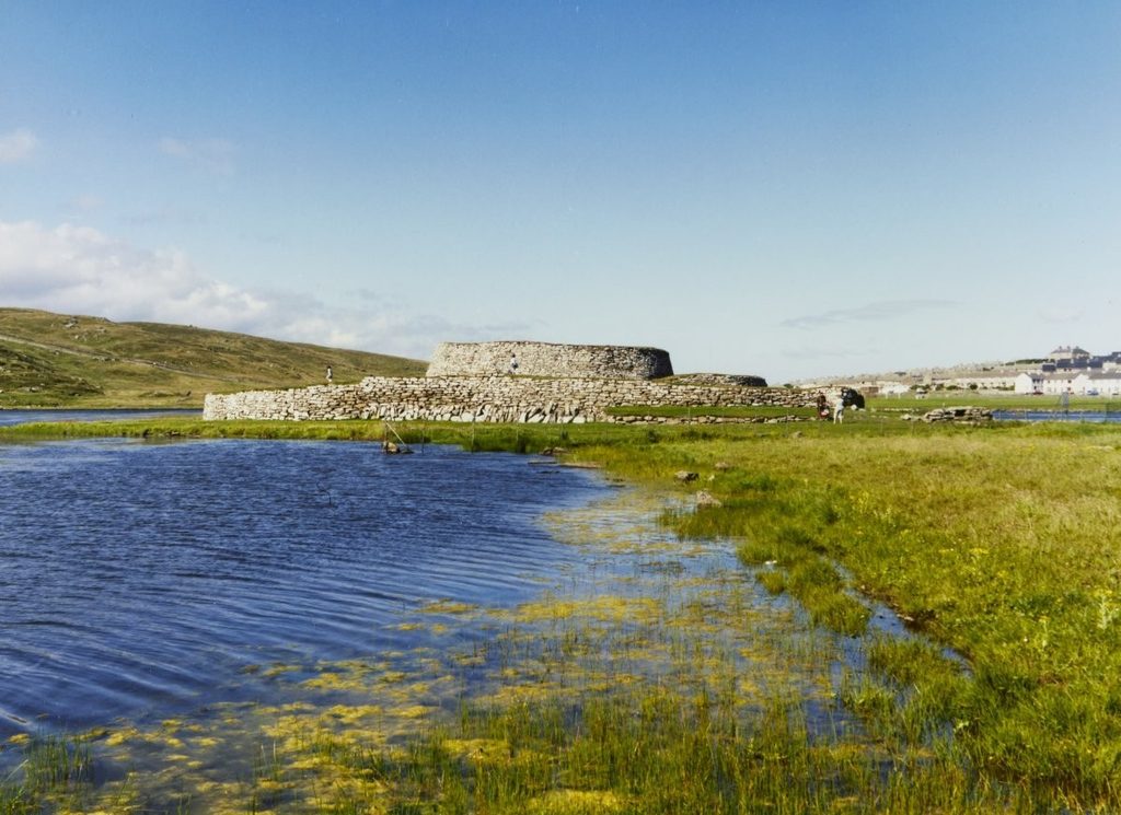 A low, drystone, structure surrounded by a lower drystone wall sit of a grassy landscape surrounded by blue water. 