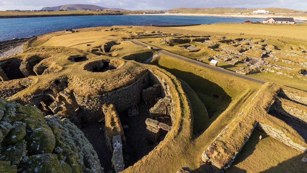 Oblique aerial view of a turf covered settlement with exposed stone walls. In the foreground, a wheelhouse, with a round external wall and spire-like internal walls is visivle. A blue body of water is visible in the background. 