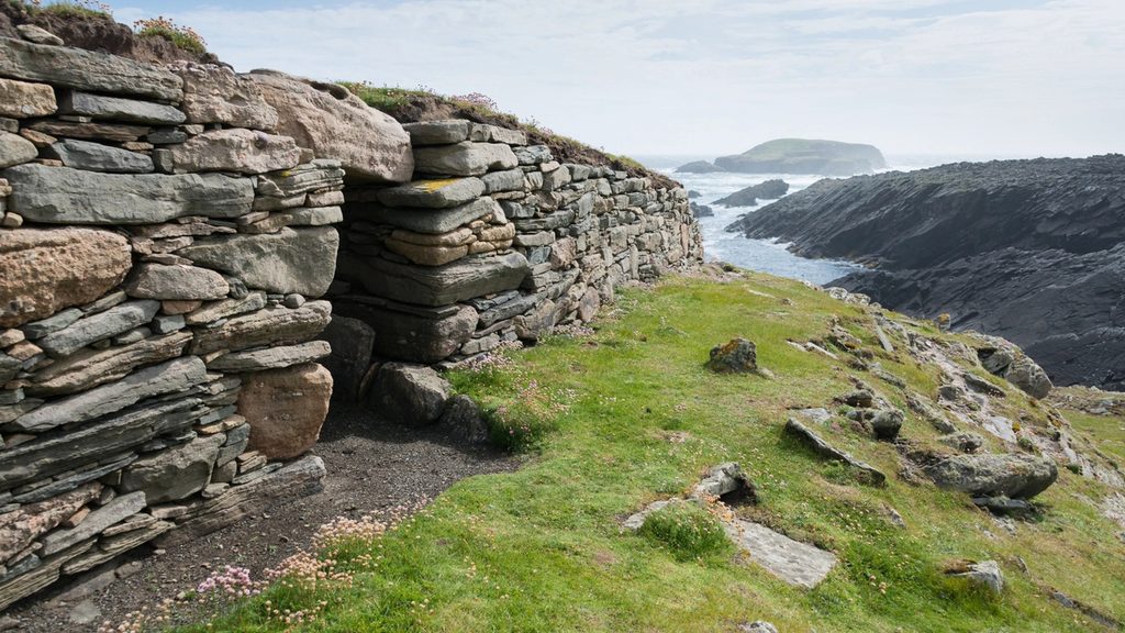 A low, drystone structure sites on a high cliff overlooking a body of water and other cliffs and islands in the background. The structure is long and rectangular, with grass growing over the top of the walls. 