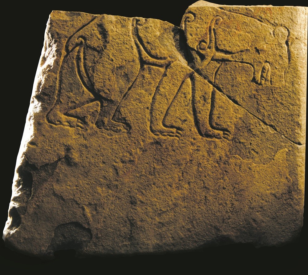 An orange-brown, square stone slabe against a black background. The top half of the stone is broken off, leaving us to assume this is the bottom of a larger stone originally. At the top of this fragment, a bear is carved into the stone. 