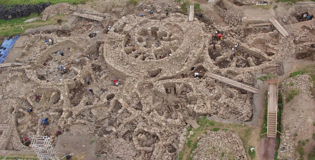 Aerial view of a very complex archaeological site, with multiple stone structures, mostly round, all arranged closely together and surrounding a much larger round building with a shamrock-shaped building in the centre. People can be seen standing and kneeling around the structures and some wooden and metal bridges and platform are placed over some of the structures to aid excavation. 