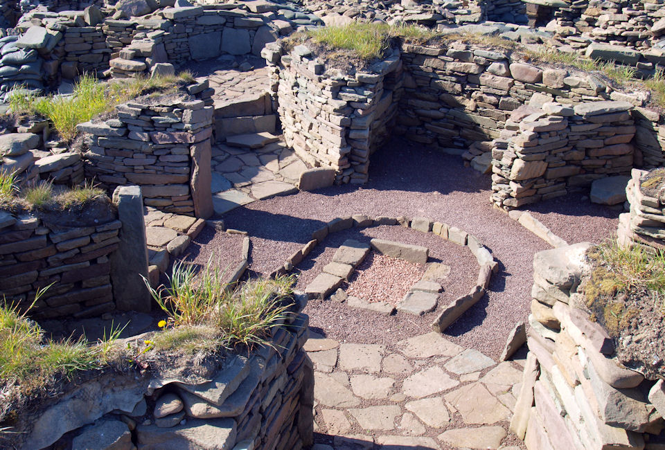 Oblique aerial photograph of a stone structure which is round, with walls resembling the spokes of a wheel. A rectangular hearth made of stone is surrounded by a semi-circle of upright stones, which looks like a fireplace or hearth. 