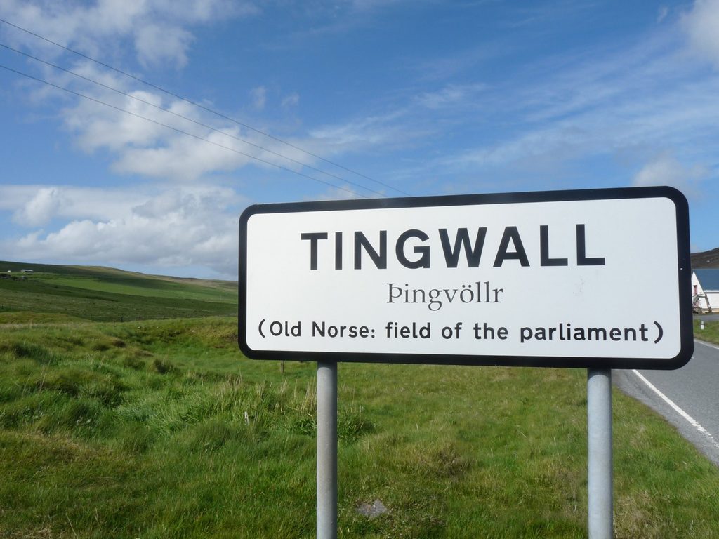 A white roadsign with black writing that says Tingwall (Old Norse: field of the parliament). The sign is set into a grassy field with a road running along its right side. 